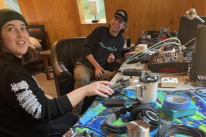 Two people laughing and interacting with.a messy table full of electronic music equipment.